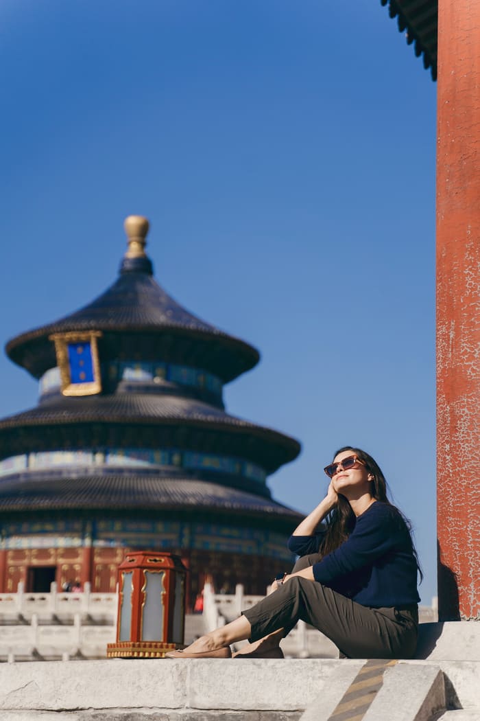 na mujer con gafas de sol sonríe sentada al aire libre, con el Templo del Cielo de Pekín