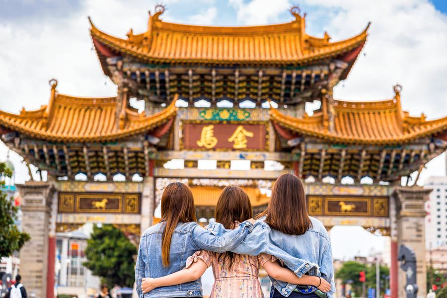 Tres mujeres, de espaldas y abrazadas, admiran la gran y decorada Puerta Jinma en Kunming, China, bajo un cielo nublado.