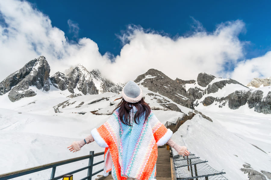 Vista trasera de una mujer con un colorido poncho tejido en tonos naranja, blanco y azul, un gorro de lana y guantes, abriendo los brazos en un sendero elevado de madera frente a una majestuosa cordillera montañosa cubierta de nieve y nubes bajo un cielo azul brillante.