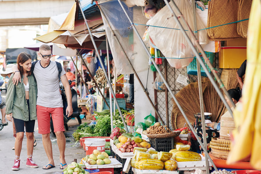 Una pareja de turistas camina abrazada por el pasillo de un mercado callejero al aire libre, pasando junto a puestos llenos de frutas, verduras frescas y diversos productos locales.