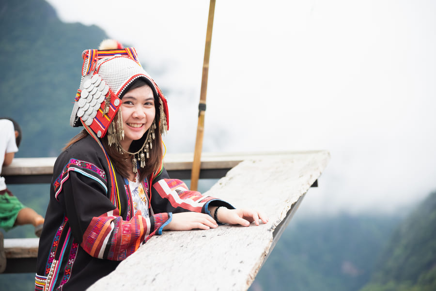Una joven sonriente que viste ropa y un elaborado tocado tradicional Akha posa apoyada en una barandilla de madera, con un paisaje de montañas neblinosas de fondo.