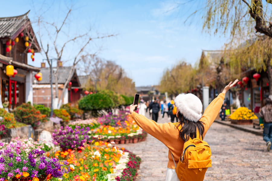 Vista trasera de una turista con mochila amarilla, suéter naranja y sombrero blanco, quien levanta un brazo con alegría mientras se toma una selfi en una calle empedrada tradicional china, rodeada de coloridos parterres de flores y arquitectura antigua.